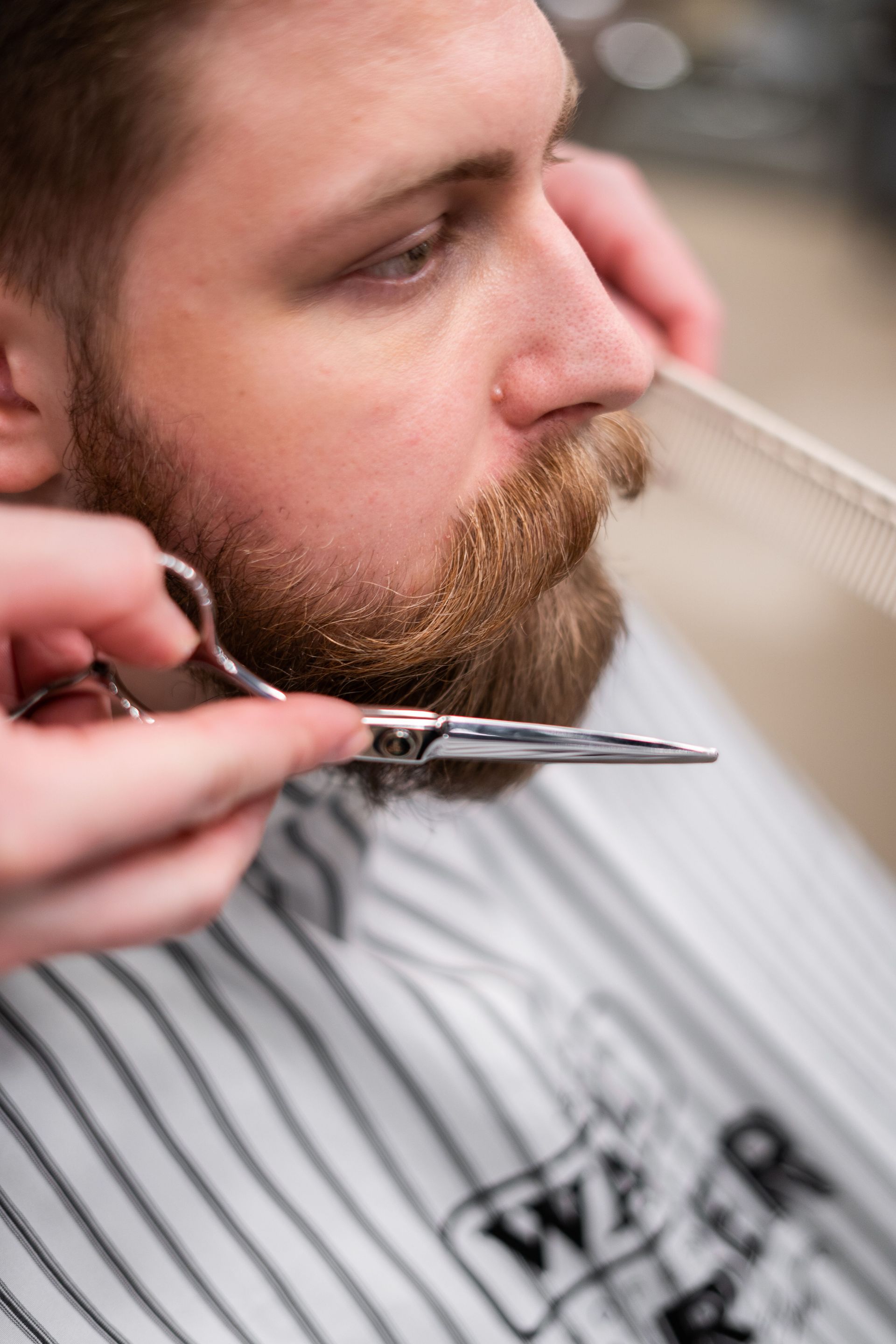 Close-up shot of a barber grooming a bearded man's beard and mustache with scissors.